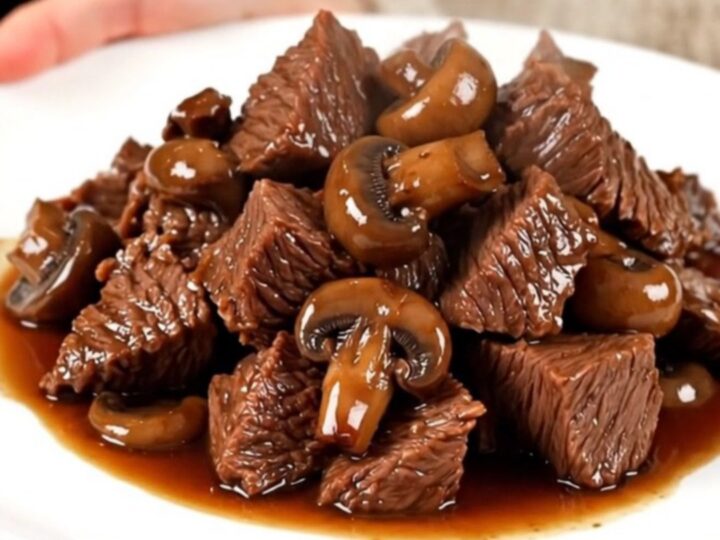 A close-up of tender, glossy braised beef chunks served with mushrooms on a white plate.