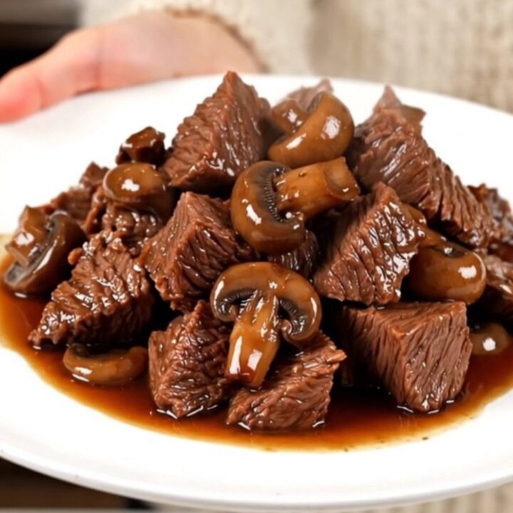 A close-up of tender, glossy braised beef chunks served with mushrooms on a white plate.