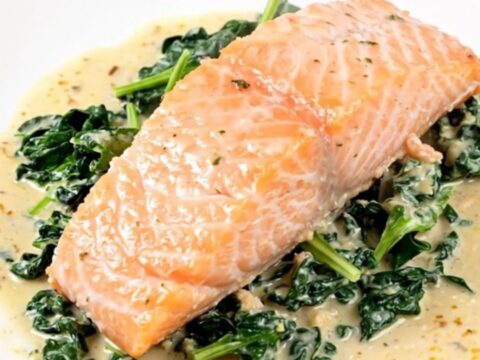 A close-up of a perfectly cooked salmon fillet covered in a creamy parmesan sauce with fresh spinach on a white plate.