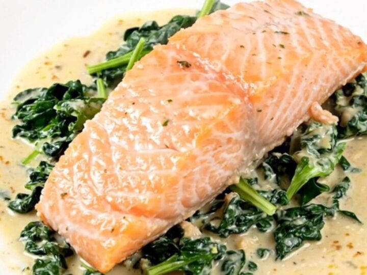 A close-up of a perfectly cooked salmon fillet covered in a creamy parmesan sauce with fresh spinach on a white plate.