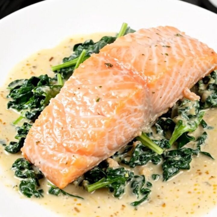 A close-up of a perfectly cooked salmon fillet covered in a creamy parmesan sauce with fresh spinach on a white plate.
