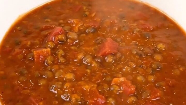 A top-down view of a black slow cooker filled with raw red lentils, diced carrots, onions, and tomato coulis.
