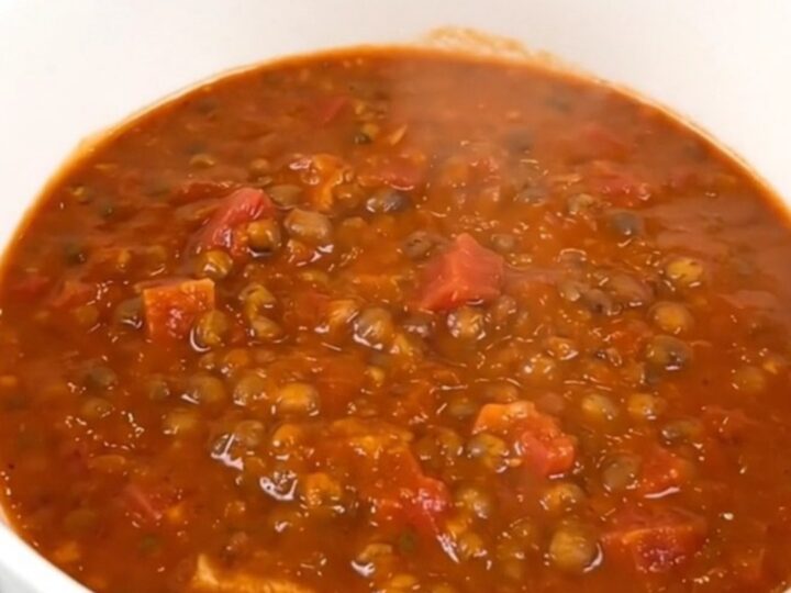 A top-down view of a black slow cooker filled with raw red lentils, diced carrots, onions, and tomato coulis.