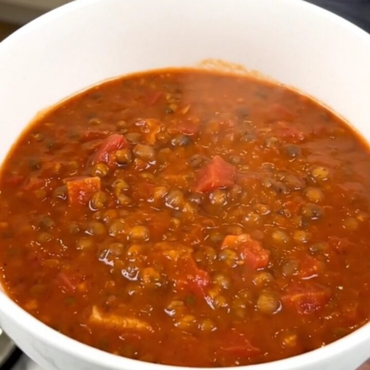 A top-down view of a black slow cooker filled with raw red lentils, diced carrots, onions, and tomato coulis.
