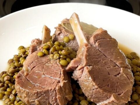 A top-down view of a black slow cooker filled with raw veal pieces, green lentils, sliced carrots, and broth.