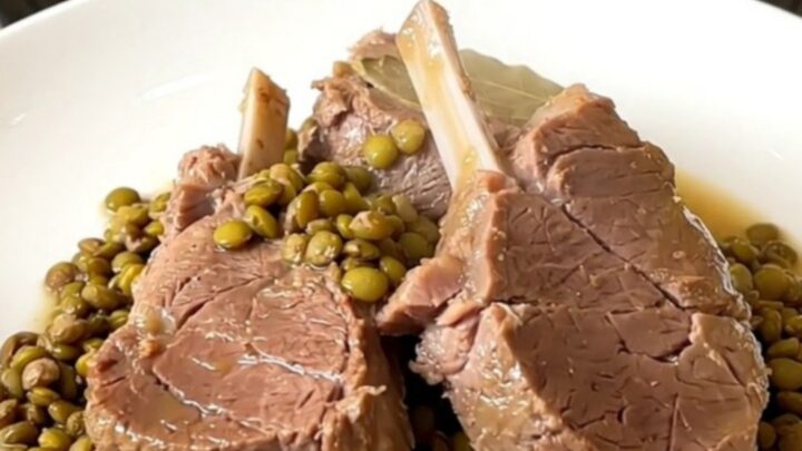 A top-down view of a black slow cooker filled with raw veal pieces, green lentils, sliced carrots, and broth.