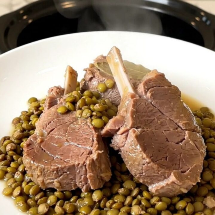 A top-down view of a black slow cooker filled with raw veal pieces, green lentils, sliced carrots, and broth.