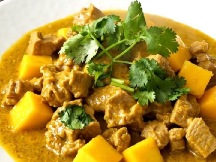 A close-up of slow cooker mango coconut pork curry served on a white plate with a garnish of cilantro.
