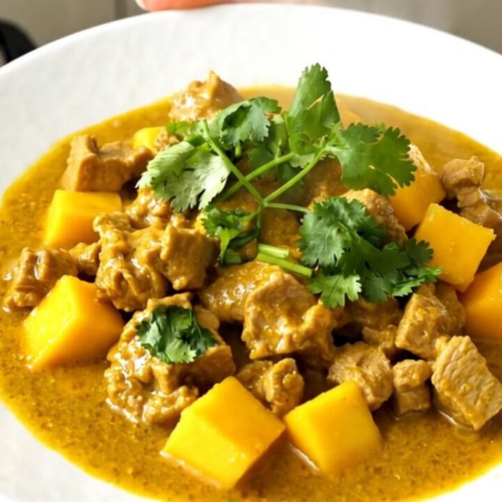 A close-up of slow cooker mango coconut pork curry served on a white plate with a garnish of cilantro.