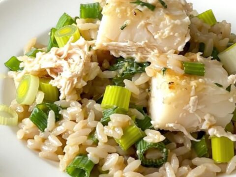 A top-down view of a black slow cooker filled with layers of fresh cod, sliced leeks, and uncooked white rice.