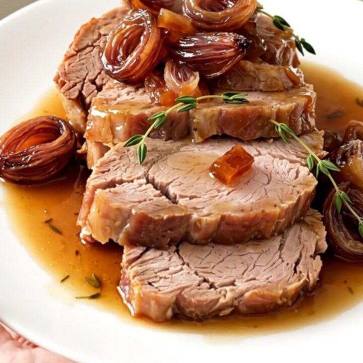 A beautifully plated slice of tender pork roast topped with glossy, caramelized shallots on a white plate.