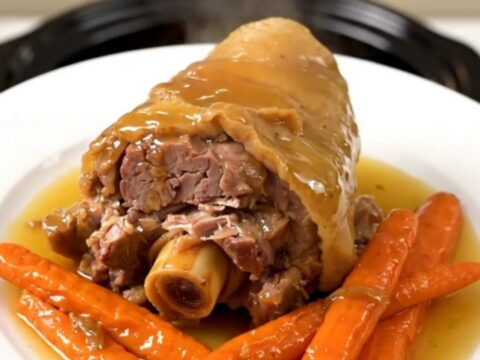 A close-up of a slow-cooked pork shank with a glossy honey glaze and tender carrots on a white plate.