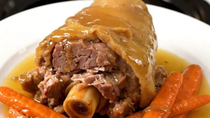 A close-up of a slow-cooked pork shank with a glossy honey glaze and tender carrots on a white plate.