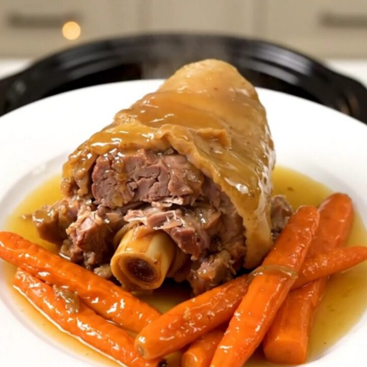 A close-up of a slow-cooked pork shank with a glossy honey glaze and tender carrots on a white plate.