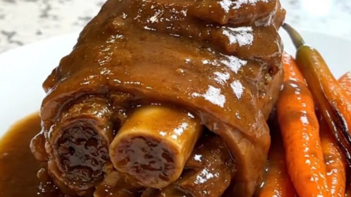 A close-up of tender, glossy lamb neck slices with cooked carrots and rich gravy on a white plate.