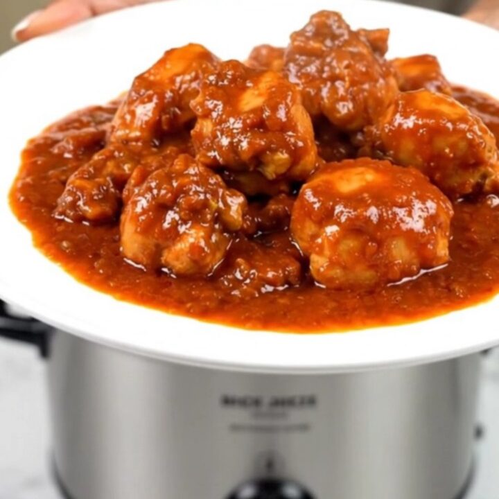 A top-down view of a black slow cooker filled with raw chicken, colorful bell peppers, onions, and tomato sauce.