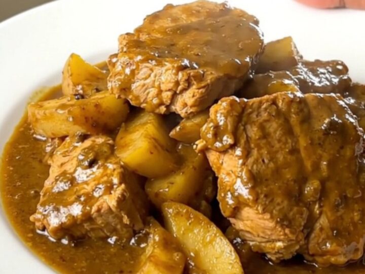 A white plate holding tender chunks of pork coated in a creamy golden curry sauce with soft, glazed apple slices.