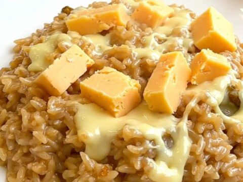 A top-down view into a black slow cooker showing raw rice, sliced onions, and chunks of Maroilles cheese.