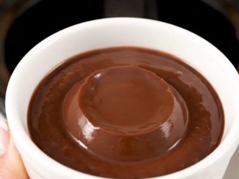 A white ceramic cup filled with thick, glossy dark chocolate pudding against a blurred kitchen background.
