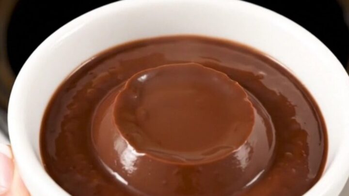 A white ceramic cup filled with thick, glossy dark chocolate pudding against a blurred kitchen background.