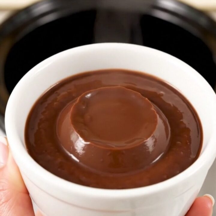 A white ceramic cup filled with thick, glossy dark chocolate pudding against a blurred kitchen background.