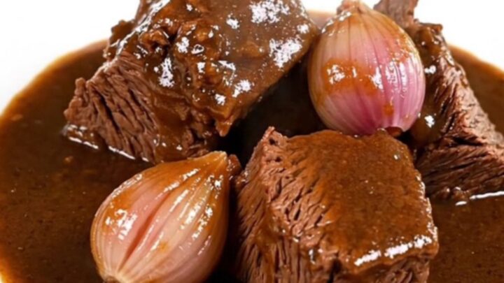 A close-up of tender beef chuck chunks in a rich, dark Madeira wine gravy with whole shallots on a white plate.