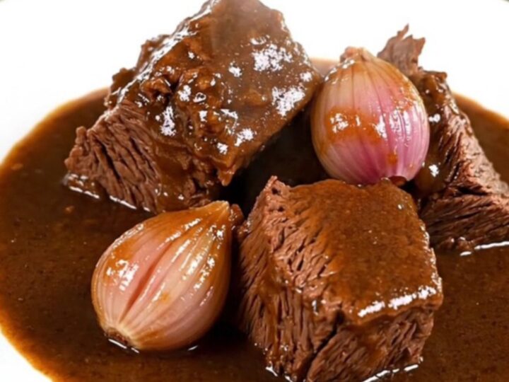 A close-up of tender beef chuck chunks in a rich, dark Madeira wine gravy with whole shallots on a white plate.