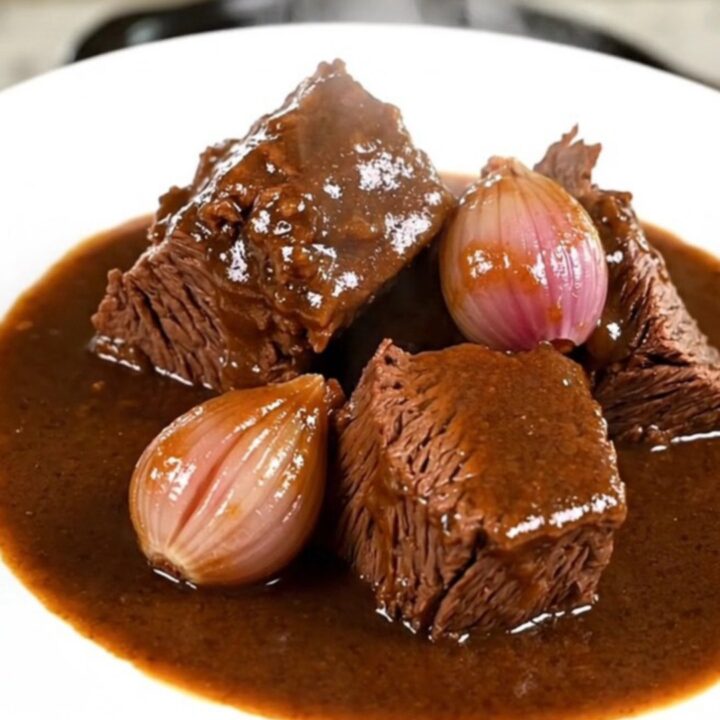 A close-up of tender beef chuck chunks in a rich, dark Madeira wine gravy with whole shallots on a white plate.