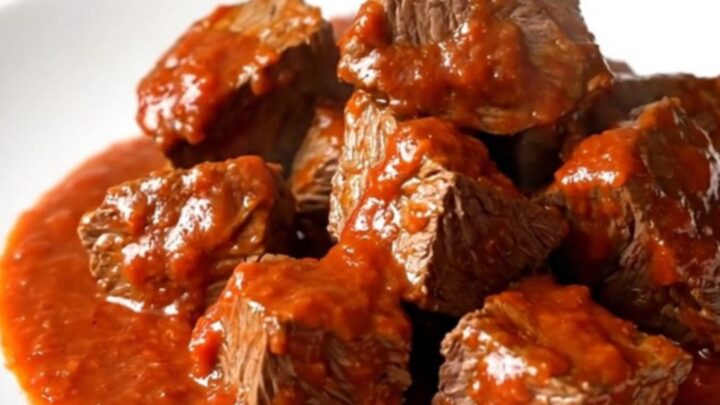 A close-up of tender, glossy beef cubes in a rich red bell pepper and tomato sauce on a white plate.
