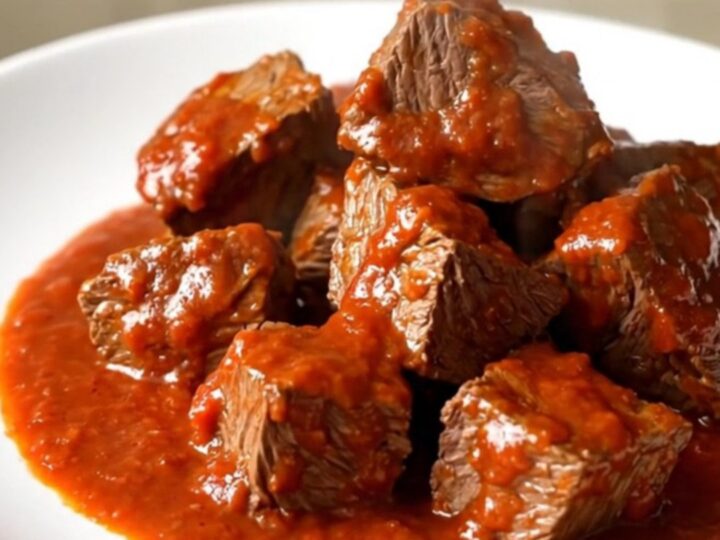 A close-up of tender, glossy beef cubes in a rich red bell pepper and tomato sauce on a white plate.