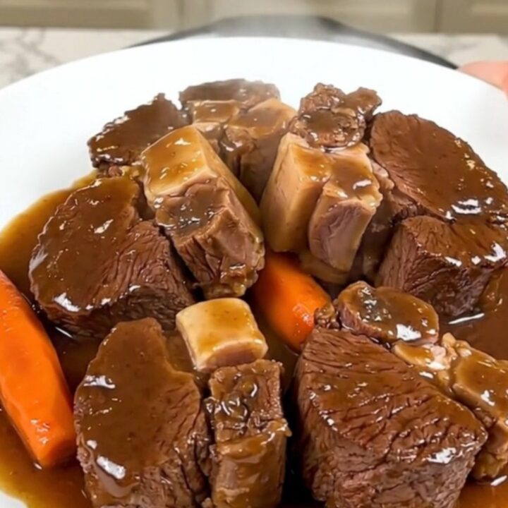 A close-up of tender, glossy beef cheeks in a rich dark sauce served with carrots on a white plate.