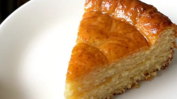 A golden-brown slice of warm apple far served on a white plate, showing a soft custard texture and apple slices.