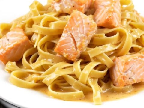 A top-down view of a black slow cooker filled with raw salmon cubes, pasta nests, and cream.