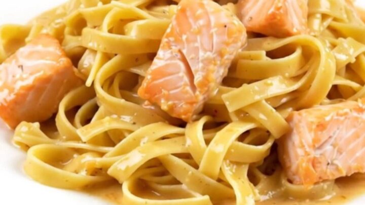 A top-down view of a black slow cooker filled with raw salmon cubes, pasta nests, and cream.