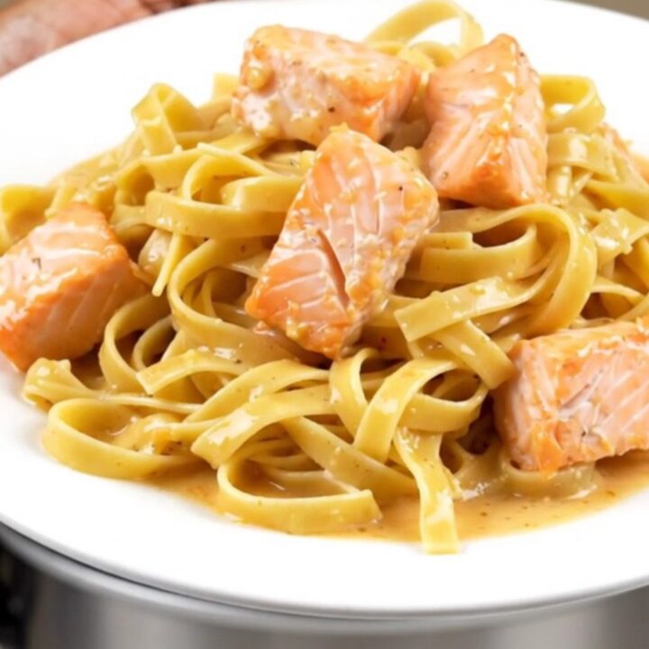 A top-down view of a black slow cooker filled with raw salmon cubes, pasta nests, and cream.