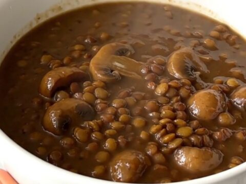 A top-down view of a black oval slow cooker filled with raw green lentils, mushrooms, and onions before cooking.