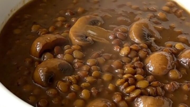 A top-down view of a black oval slow cooker filled with raw green lentils, mushrooms, and onions before cooking.