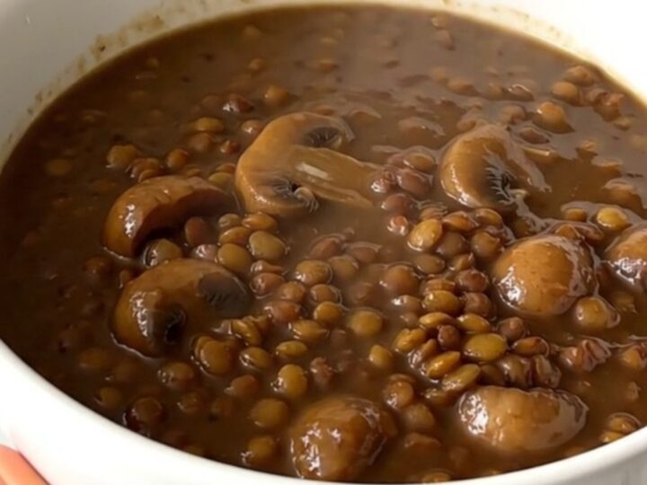 A top-down view of a black oval slow cooker filled with raw green lentils, mushrooms, and onions before cooking.