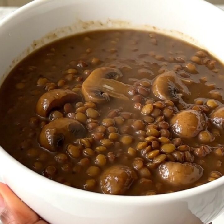 A top-down view of a black oval slow cooker filled with raw green lentils, mushrooms, and onions before cooking.