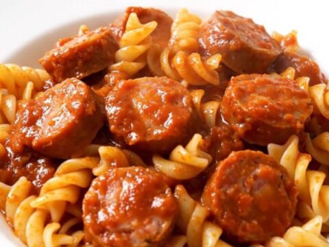 A top-down view of a black slow cooker filled with sliced sausages, onions, and tomato sauce before cooking.