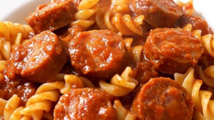 A top-down view of a black slow cooker filled with sliced sausages, onions, and tomato sauce before cooking.
