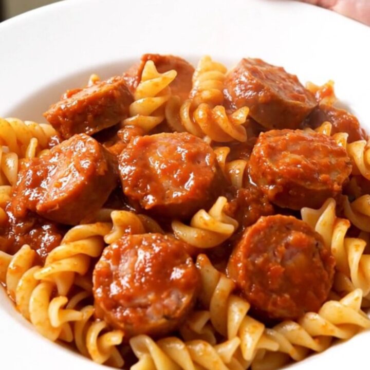 A top-down view of a black slow cooker filled with sliced sausages, onions, and tomato sauce before cooking.