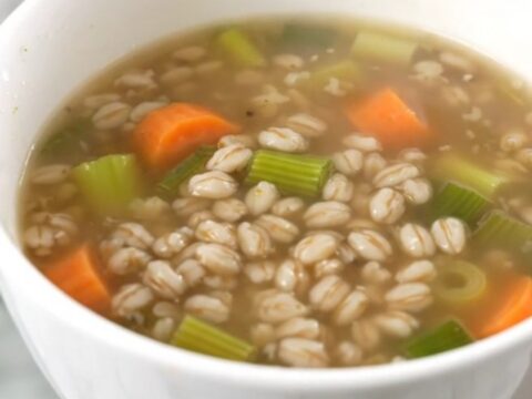 A steaming white bowl filled with thick vegetable and pearl barley soup on a kitchen counter.