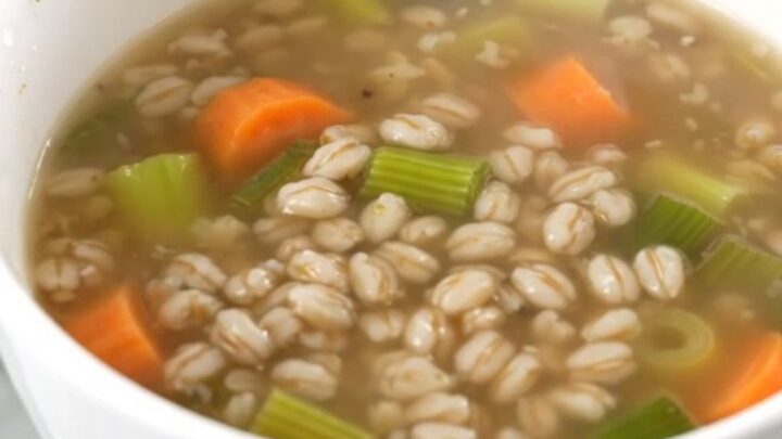 A steaming white bowl filled with thick vegetable and pearl barley soup on a kitchen counter.
