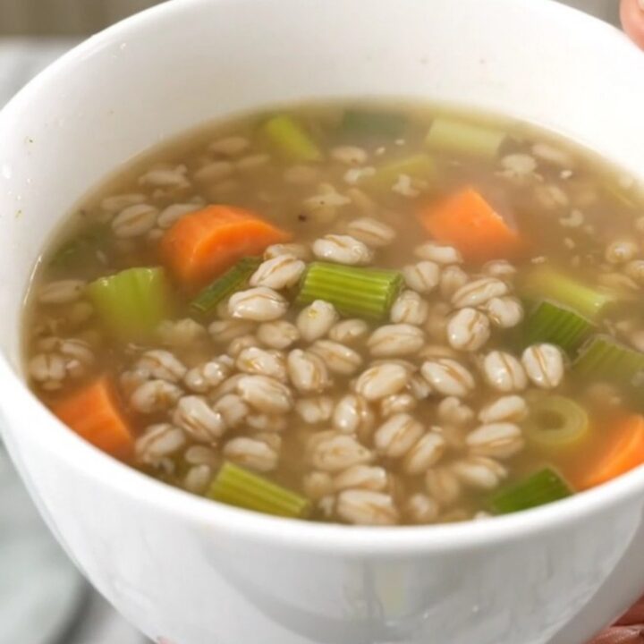A steaming white bowl filled with thick vegetable and pearl barley soup on a kitchen counter.