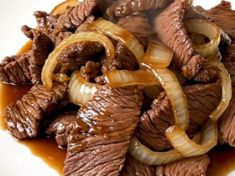 A top-down view of raw beef, sliced onions, and garlic in a black slow cooker bowl.