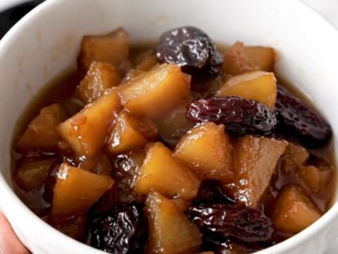 A white ceramic bowl filled with warm, glossy slow-cooked apple and prune compote in a rustic kitchen.