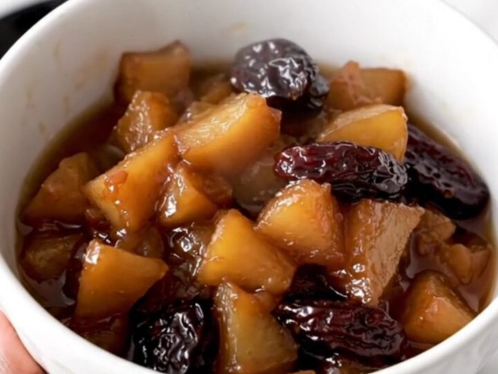 A white ceramic bowl filled with warm, glossy slow-cooked apple and prune compote in a rustic kitchen.