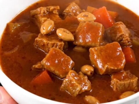 A close-up of a steaming bowl of pork peanut stew with vibrant carrots and a rich, glossy sauce.