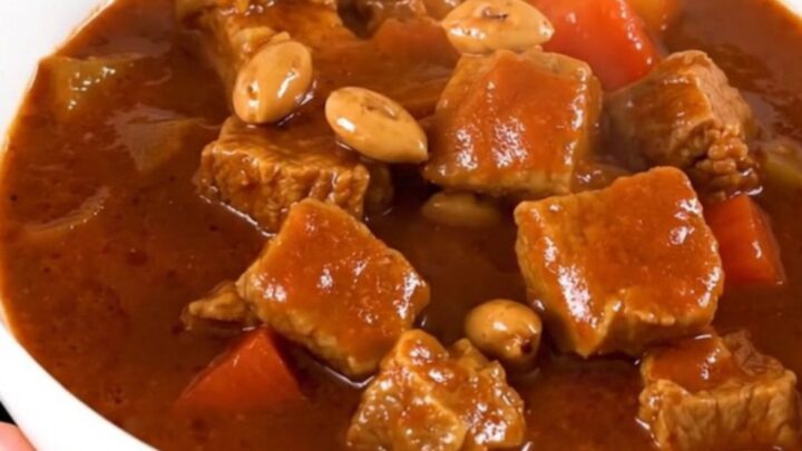 A close-up of a steaming bowl of pork peanut stew with vibrant carrots and a rich, glossy sauce.